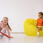 Two children enjoying playtime indoors with a hula hoop and bean bag, full of joy and fun.