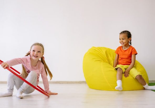 Two children enjoying playtime indoors with a hula hoop and bean bag, full of joy and fun.